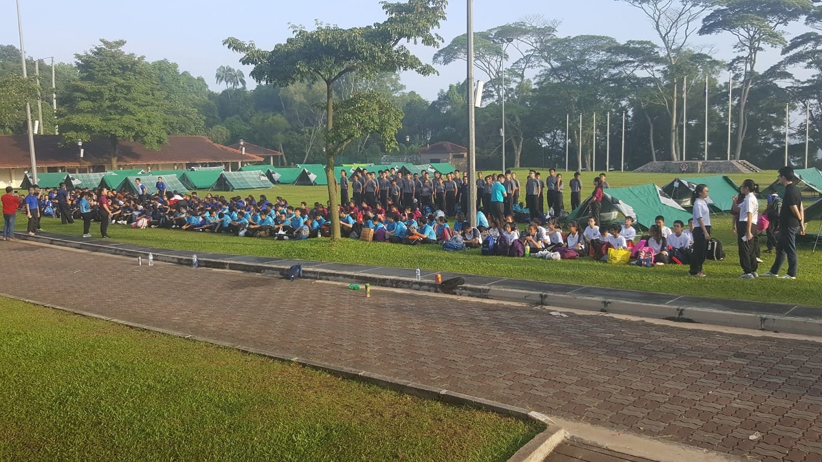 A large group of students gathers outdoors on a grassy field, sitting and standing in organized rows beside a paved walkway. Many green tents are set up behind them, forming a camp area. 